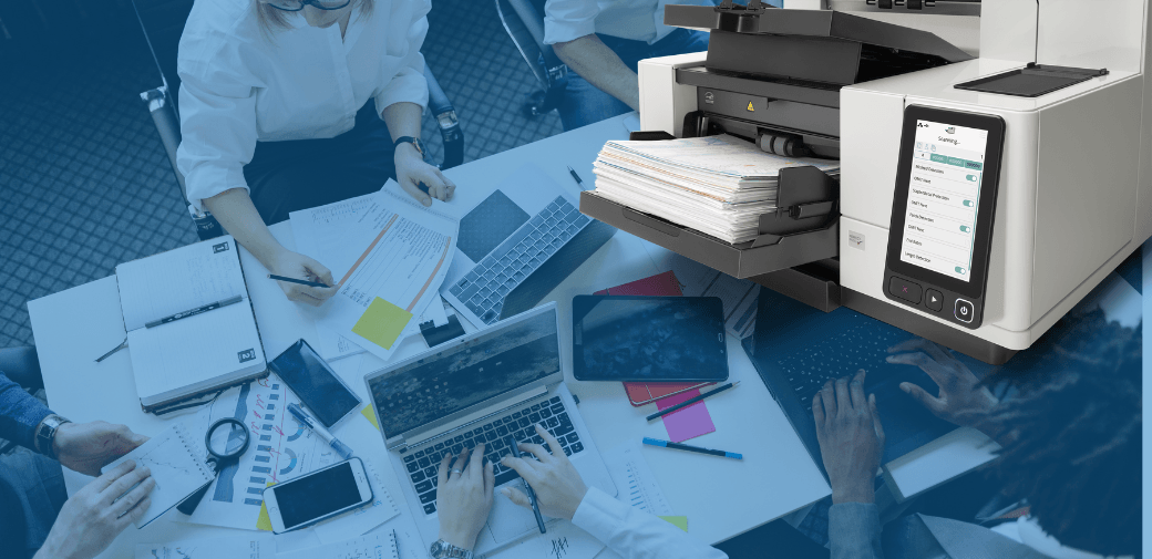 A high-speed document scanner sits on a desk next to a group of professionals working with laptops, tablets, and paperwork in a modern off